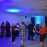 18 Grupa osób tańczących w jasno oświetlonej sali z niebieskim światłem. Tło z bannerem Mazowsze. Ubrania w odcieniach czerwieni i czerni.