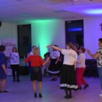 19 Grupa osób tańczy w jasnym pomieszczeniu z niebieskim oświetleniem. Kobieta w centrum nosi biały T-shirt z napisem. W tle banery i krzyż na ścianie.