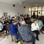 Grupa ludzi siedzi na krzesłach w jasnym pomieszczeniu z białymi ścianami. Uczestnicy rozmawiają między sobą, niektórzy trzymają notatki. W tle jest plakat i drewniane drzwi.