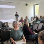 Grupa seniorów siedzi w sali szkoleniowej. Na ekranie pokazane są instrukcje ćwiczenia Rysunek w ciemno. Ujęcie przedstawia różnorodne interakcje i wymianę zdań.