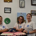 20 Trzy kobiety w białych koszulkach siedzą przy stole na tle obrazów. Na ścianie zielony znak Uniwersytet Dobrego Wieku. Na stole różowe wstążki i ulotki.