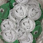 Kosz pełen okrągłych przypinek. Przypinki są w kolorach białym z czarnym tekstem i zielonym z czarnym obrazem. Przedstawiają rysunek i teksty upamiętniające oraz logo.