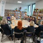 Na obrazie widać grupę ludzi siedzących wokół drewnianego stołu w bibliotece. Kobieta prowadzi prezentację, a w tle są regały z książkami. Ściany są jasne, z dużymi oknami i zielonymi roślinami.