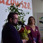 3 Osoba w okularach mówi do mikrofonu, trzymając bukiet. Obok stoi kobieta. W tle napisy i roślina na białej ścianie.