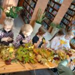 Dzieci przy stole pełnym kolorowych liści, w bibliotece z książkami na regałach. Słoneczne światło pada na wnętrze.