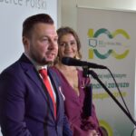 4 Mężczyzna w garniturze i kobieta w sukni stoją przy mikrofonie. Tło z plakatami w jasnych kolorach i napisami. Jasne oświetlenie na scenie.