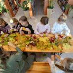 Dzieci siedzą przy drewnianym stole, układając kolorowe liście. Tło to wnętrze biblioteki z książkami i roślinami w doniczkach. Atmosfera jest przytulna i jesienna.