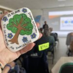 Ręka trzyma pomalowany kamień z drzewem i napisem otoczony kwiatami. W tle widać salę konferencyjną z prezentacją. Kolory to zieleń, błękit i brąz.