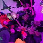 Grupa dzieci w kostiumach halloweenowych siedzi w kręgu, ręce wyciągnięte do środka. Tło jest fioletowe. Na podłodze drewno.