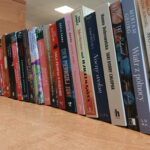 Na zdjęciu widać rząd książek ustawionych pionowo na drewnianym blacie. Okładki są kolorowe, a w tle znajduje się jasna ściana. Oświetlenie jest jasne, prawdopodobnie sztuczne.