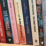 Zdjęcie przedstawia rząd książek na półce, z kolorowymi grzbietami w odcieniach czerwieni, niebieskiego i beżu. Tło jest neutralne, drewniane. Grzbiety zawierają tytuły i nazwiska autorów.