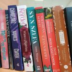 Półka z kolorowymi książkami; różne tytuły, głównie w pionie. Tło jasne, dominują odcienie czerwieni i fioletu.