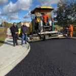 Na obrazie jest maszyna do układania asfaltu otoczona przez kilku pracowników w odzieży odblaskowej. Tło stanowi niebieskie niebo z białymi chmurami oraz drzewa. Na chodniku stoją dwaj mężczyźni w ciemnych kurtkach.