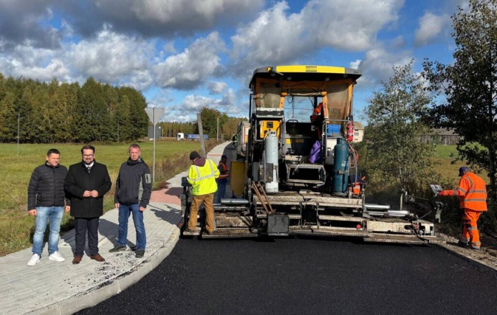 Na zdjęciu roboty drogowe z maszyną do asfaltu i grupą ludzi. Tło to zielone drzewa i błękitne niebo z chmurami. Robotnicy w jaskrawych kamizelkach pracują obok maszyny.