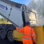 Na zdjęciu widać mężczyznę w pomarańczowym stroju, obok ciężarówki z napisem Schmitz Cargobull. Pojazd ma szare koła, ładunek wysypuje się do żółtej maszyny. W tle jest niebo i zieleń dr