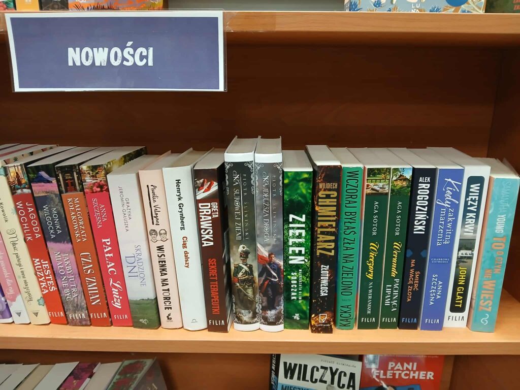 Półka z nowościami w księgarni, pełna kolorowych grzbietów książek. Tło to drewniana półka. Widoczny napis NOWOŚCI.