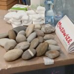 Na stole leżą kolorowe kamienie i białe mydła. Obok stoją butelki wody. W tle są markery i kartka z napisem.