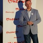 Mężczyzna w jasnoszarej marynarce stoi przed białym tłem z napisem Mazowsze w czerwieni. Tło jest jednolite, a napis wielokrotny. Atmosfera jest oficjalna, z zacienioną sylwetką.