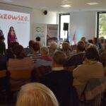 6 Spotkanie w audytorium, na scenie widoczni są mówca i prezenterka. Jasne tło z logo Mazowsze i szare krzesła. Uczestnicy siedzą tyłem do obiektywu.