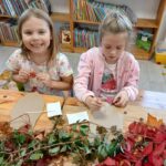 Na zdjęciu dwie dziewczynki przy stole dekorują liśćmi. Stół pokryty jest liśćmi w odcieniach czerwieni i zieleni. W tle półki z książkami.