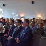 7 Grupa osób siedzi w jasnym pomieszczeniu. Mężczyźni w garniturach mają różowe wstążki. Ściany są jasnoszare z lampami.