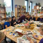 Grupa osób siedzi przy stole z przekąskami i materiałami artystycznymi. W tle regały z książkami i dekoracyjne rośliny. Kolory są jasne z dominacją drewna i zieleni.