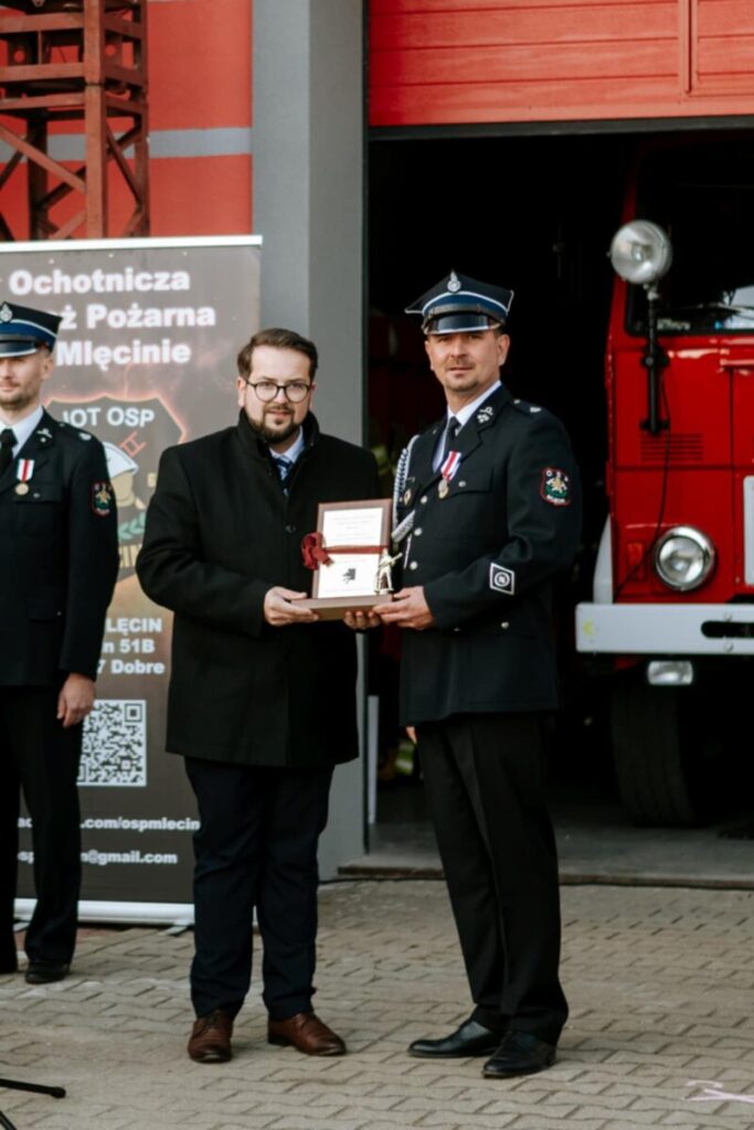 Dwóch mężczyzn stoi na tle czerwonego wozu strażackiego. Jeden wręcza drugiemu dyplom, obaj mają uroczyste stroje. Po lewej widnieje baner Ochotniczej Straży Pożarnej.