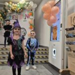 Dwoje dzieci w kostiumach na Halloween, stoją przed dekoracją z balonów. Tło zdobi napis Dzień Dyni i kolorowe balony. Na prawo widoczna jest elektroniczna tablica z informacjami.