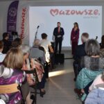 8 Obraz przedstawia salę z ludźmi, dwoje mówców na scenie, tło z logo Mazowsza. Dominuje jasne oświetlenie i ciepłe kolory.