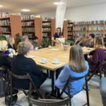 Na zdjęciu widać grupę osób siedzących przy stole w bibliotece. W tle półki z książkami, na stole filiżanki. Osoba stojąca prowadzi dyskusję.