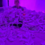 Na zdjęciu jest osoba otoczona mnóstwem papieru. Oświetlenie w kolorze fioletowym dominuje cały obraz. Tło jest jednolite, co podkreśla centralną scenę.