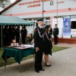 Ceremonia przed strażnicą OSP w Mileńcu, ludzie w mundurach. Na pierwszym planie stół z nagrodami, w tle namiot i budynek.