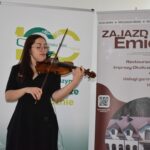 9 Kobieta gra na skrzypcach, stojąc przed dwoma banerami. Ubrana jest na czarno, tło jest jasne z tekstem i grafiką. Na jednym banerze widoczna jest reklama hotelu.