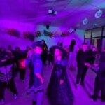 Dzieci w kostiumach na przyjęciu halloweenowym, oświetlone fioletowym światłem. Tło: girlanda balonowa, wiszące dynie. Sala pełna uczestników w strojach.