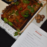 Na białym obrusie leży prostokątne ciasto ozdobione ziołami i warzywami. Obok są orzechy, jabłka i miska z ogórkami. W prawym dolnym rogu widoczny jest dokument z logo Mazowsze.