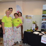 Pięć osób stoi obok stoiska z ulotkami i jedzeniem. Dwie kobiety w zielonych koszulkach mają kwieciste spódnice. Tło jasne, z plakatem promującym Gminę Dobre.