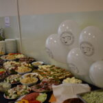 Na stole z ciemnym obrusem ułożono różnorodne sałatki i kanapki. Obok stoją białe balony z nadrukiem oraz stos talerzy. Tło jest jasne, dekoracje są minimalistyczne.