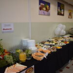 Stół szwedzki z daniami na czarnym obrusie. Tło beżowe z obrazami, balony i zastawa. Posiłki w kolorach warzyw i owoców.