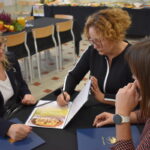 Trzy kobiety siedzą przy stole i przeglądają dokumenty. Tło wypełniają krzesła i stół z czarnym obrusem. Kolory: czarny, biały, jasnobrązowy.