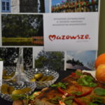 Stół z potrawami regionalnymi: ziemniaki, kiszone ogórki, ozdoby z ziół. W tle plansza Mazowsze. Jabłka i dynia obok.