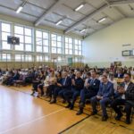 Sala gimnastyczna z jasnym ścianami i oknami, wypełniona ludźmi na krzesłach. Na podłodze jasne panele, kosz do koszykówki w tle. Wszyscy obecni siedzą i patrzą w jednym kierunku.