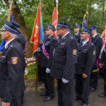 Grupa strażaków w mundurach stoi z kolorowymi sztandarami. Tło to zielone drzewa. Ujęcie z uroczystości plenerowej.