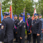Grupa mężczyzn w mundurach strażackich z flagami stoi na tle zieleni. Mają na sobie ciemne uniformy z niebieskimi elementami. Tło stanowią drzewa z gęstymi liśćmi.