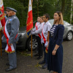 Grupa osób w odświętnych strojach stoi w szeregu z flagą Polski. Tło to zielony las. Przeważają kolory czerwony, biały i niebieski.