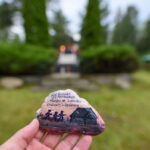 Ktoś trzyma pomalowany kamień z napisem i rysunkami. W tle rozmyte polskie flagi i zielone krzewy. Kolory: zielony, biały, czerwony.