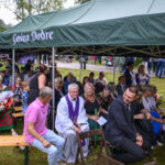 Grupa ludzi pod zielonym namiotem z napisem Gmina Dobre, siedzą na ławkach, tło stanowi zieleń drzew i trawa.