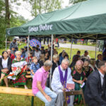 Grupa ludzi siedzi pod zielonym namiotem z napisem Gmina Dobre na tle drzew. Mają na sobie kolorowe ubrania. Pogoda pochmurna.