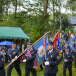Grupa strażaków w paradzie niesie kolorowe flagi. Mundury są granatowe, a tło zielone od drzew. Widać namiot i widzów.