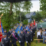 Grupa strażaków w mundurach niesie kolorowe sztandary, stojąc obok zielonego namiotu. W tle drzewa i dom. Uroczystość na świeżym powietrzu.