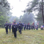 Grupa ludzi w mundurach maszeruje w deszczu z flagami w rękach. Tło to zielone drzewa i trawa. Obok stoi osoba przy masztu z biało-czerwoną flagą.